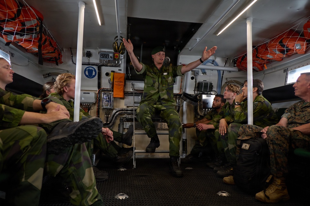 Military personnel are gathered inside what appears to be a naval vessel's interior compartment. A soldier wearing a green beret is standing/sitting centrally and appears to be briefing or instructing the group, with arms raised in an explanatory gesture. The group includes personnel in Swedish-pattern camouflage uniforms and at least one individual in US Marine Corps digital camouflage.