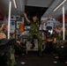 Military personnel are gathered inside what appears to be a naval vessel's interior compartment. A soldier wearing a green beret is standing/sitting centrally and appears to be briefing or instructing the group, with arms raised in an explanatory gesture. The group includes personnel in Swedish-pattern camouflage uniforms and at least one individual in US Marine Corps digital camouflage.