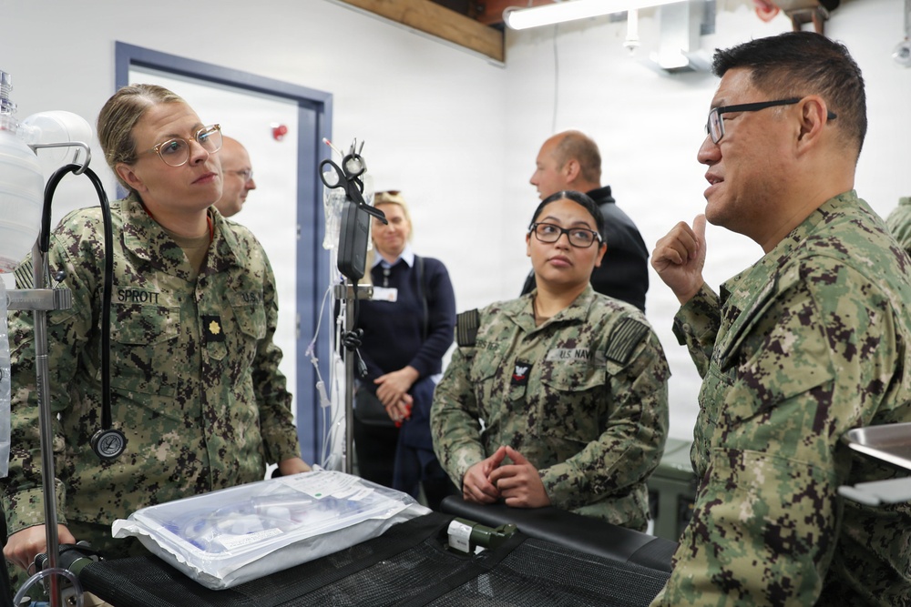 Three U.S. Navy personnel in NWU Type III camouflage uniforms are engaged in a discussion around what appears to be a medical examination or treatment table in a clinical or field medical facility. The individual on the left, identified by name tape as 'SPROTT,' appears to be listening attentively while the individual on the right gestures as if explaining something. Additional personnel in civilian and military attire are visible in the background.
