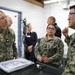 Three U.S. Navy personnel in NWU Type III camouflage uniforms are engaged in a discussion around what appears to be a medical examination or treatment table in a clinical or field medical facility. The individual on the left, identified by name tape as 'SPROTT,' appears to be listening attentively while the individual on the right gestures as if explaining something. Additional personnel in civilian and military attire are visible in the background.