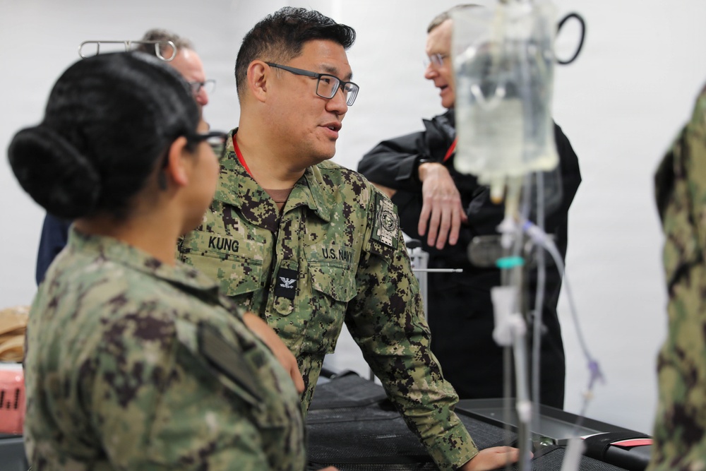 U.S. Navy personnel in digital camouflage uniforms are gathered around what appears to be a medical station or treatment area. A central figure identified by name tape as 'KUNG' with U.S. Navy insignia appears to be briefing or instructing colleagues. An IV bag and medical tubing are visible in the foreground, suggesting a medical training or operational medical environment.
