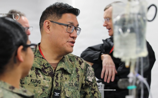 U.S. Navy personnel in digital camouflage uniforms are gathered around what appears to be a medical station or treatment area. A central figure identified by name tape as 'KUNG' with U.S. Navy insignia appears to be briefing or instructing colleagues. An IV bag and medical tubing are visible in the foreground, suggesting a medical training or operational medical environment.