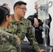 U.S. Navy personnel in digital camouflage uniforms are gathered around what appears to be a medical station or treatment area. A central figure identified by name tape as 'KUNG' with U.S. Navy insignia appears to be briefing or instructing colleagues. An IV bag and medical tubing are visible in the foreground, suggesting a medical training or operational medical environment.