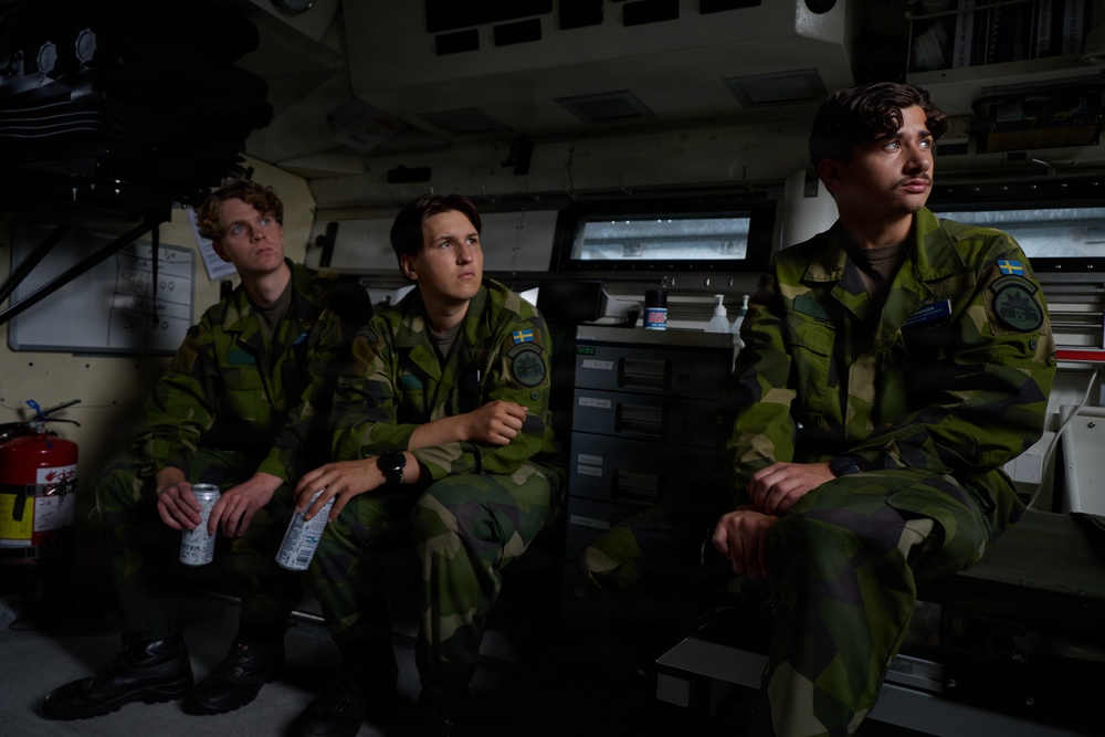 Three young soldiers in Swedish military camouflage uniforms are seated inside what appears to be a military vehicle or command post interior. They are wearing identical M90 pattern camouflage uniforms with visible Swedish flag patches on their shoulders. The setting appears to be the interior of an armored personnel carrier or similar military vehicle.