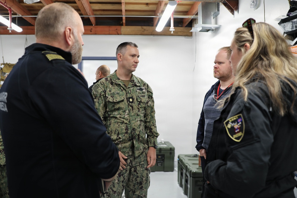 A group of four individuals are engaged in a discussion inside what appears to be a military or emergency management facility. One person is wearing U.S. Navy camouflage (NWU Type III) with the name tape 'BUCCINE' and a gold oak leaf insignia indicating the rank of Lieutenant Commander. The others appear to include law enforcement or emergency management personnel, identifiable by patches and tactical clothing.