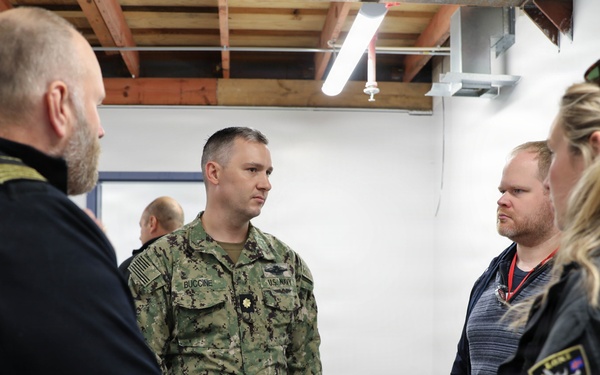 A group of four individuals are engaged in a discussion inside what appears to be a military or emergency management facility. One person is wearing U.S. Navy camouflage (NWU Type III) with the name tape 'BUCCINE' and a gold oak leaf insignia indicating the rank of Lieutenant Commander. The others appear to include law enforcement or emergency management personnel, identifiable by patches and tactical clothing.