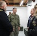A group of four individuals are engaged in a discussion inside what appears to be a military or emergency management facility. One person is wearing U.S. Navy camouflage (NWU Type III) with the name tape 'BUCCINE' and a gold oak leaf insignia indicating the rank of Lieutenant Commander. The others appear to include law enforcement or emergency management personnel, identifiable by patches and tactical clothing.