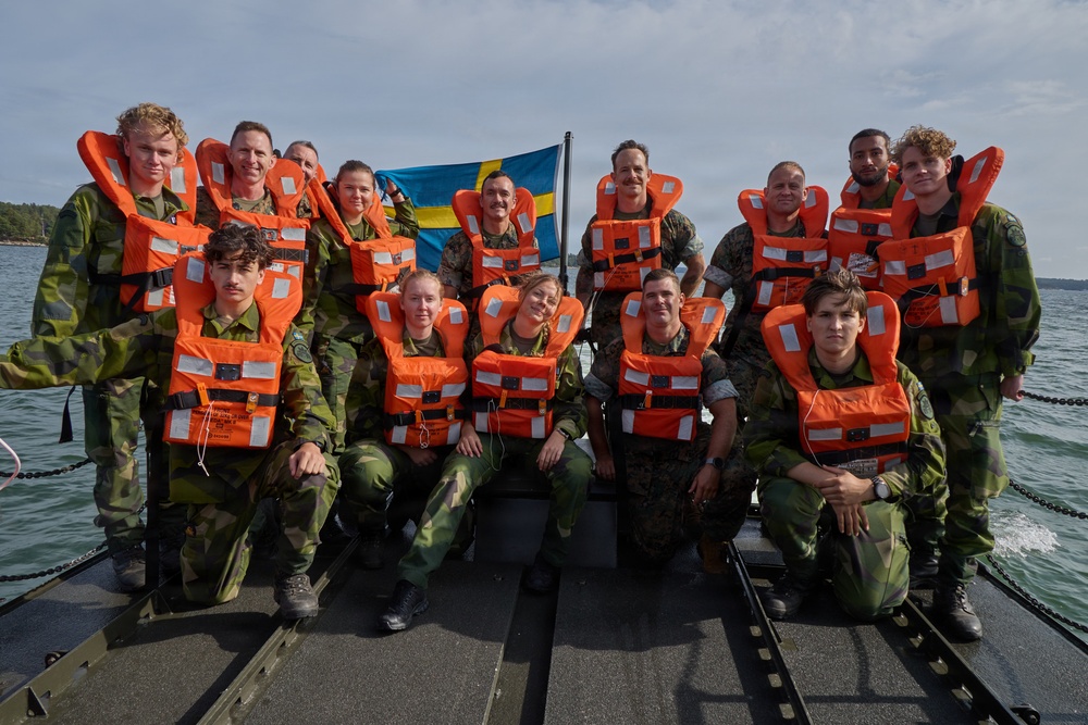 A group of approximately 15 military personnel in camouflage uniforms are posing for a group photo on a military watercraft. All individuals are wearing bright orange life vests, and a Swedish flag is prominently displayed in the background. The setting appears to be a coastal or archipelago environment with calm water and forested shoreline visible.