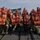 A group of approximately 15 military personnel in camouflage uniforms are posing for a group photo on a military watercraft. All individuals are wearing bright orange life vests, and a Swedish flag is prominently displayed in the background. The setting appears to be a coastal or archipelago environment with calm water and forested shoreline visible.