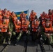 A group of approximately 15 military personnel in camouflage uniforms are posing for a group photo on a military watercraft. All individuals are wearing bright orange life vests, and a Swedish flag is prominently displayed in the background. The setting appears to be a coastal or archipelago environment with calm water and forested shoreline visible.