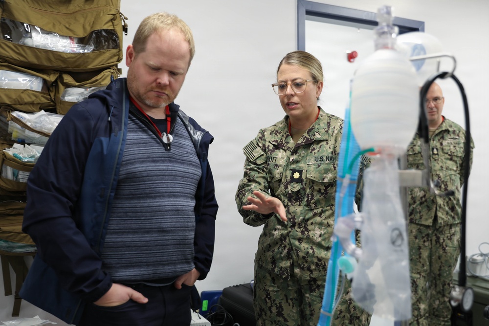 A U.S. Navy officer in NWU Type III camouflage uniform is briefing a civilian on medical equipment, likely an IV or respiratory apparatus. The setting appears to be a medical training or demonstration room with tactical medical supply bags visible in the background. A second uniformed Navy personnel is visible in the background observing the interaction.
