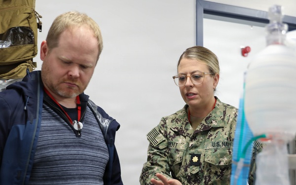 A U.S. Navy officer in NWU Type III camouflage uniform is briefing a civilian on medical equipment, likely an IV or respiratory apparatus. The setting appears to be a medical training or demonstration room with tactical medical supply bags visible in the background. A second uniformed Navy personnel is visible in the background observing the interaction.