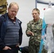 A U.S. Navy officer in NWU Type III camouflage uniform is briefing a civilian on medical equipment, likely an IV or respiratory apparatus. The setting appears to be a medical training or demonstration room with tactical medical supply bags visible in the background. A second uniformed Navy personnel is visible in the background observing the interaction.