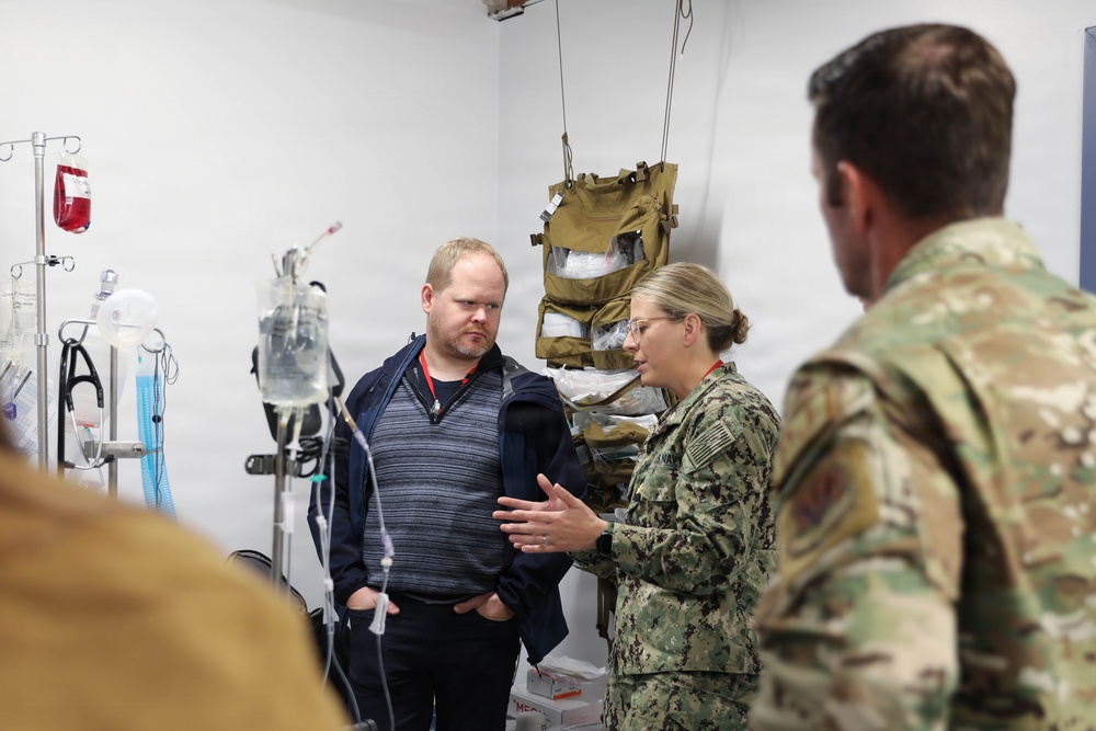 A military medical briefing or tour is taking place in what appears to be a field medical treatment room or aid station. A female Navy service member in NWU Type III (digital camouflage) uniform is speaking to a civilian male wearing a red lanyard badge and a soldier in Army OCP (Operational Camouflage Pattern) uniform. Medical equipment and tactical gear are visible in the background.