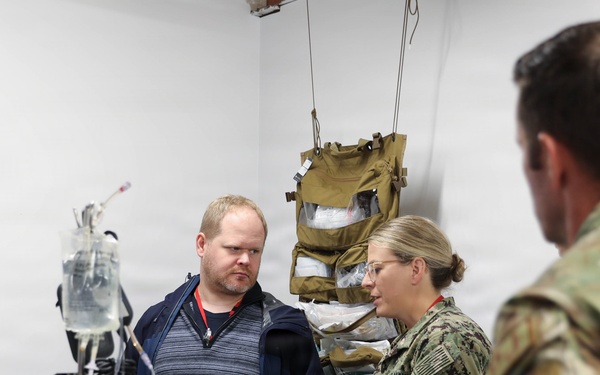 A military medical briefing or tour is taking place in what appears to be a field medical treatment room or aid station. A female Navy service member in NWU Type III (digital camouflage) uniform is speaking to a civilian male wearing a red lanyard badge and a soldier in Army OCP (Operational Camouflage Pattern) uniform. Medical equipment and tactical gear are visible in the background.