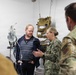 A military medical briefing or tour is taking place in what appears to be a field medical treatment room or aid station. A female Navy service member in NWU Type III (digital camouflage) uniform is speaking to a civilian male wearing a red lanyard badge and a soldier in Army OCP (Operational Camouflage Pattern) uniform. Medical equipment and tactical gear are visible in the background.
