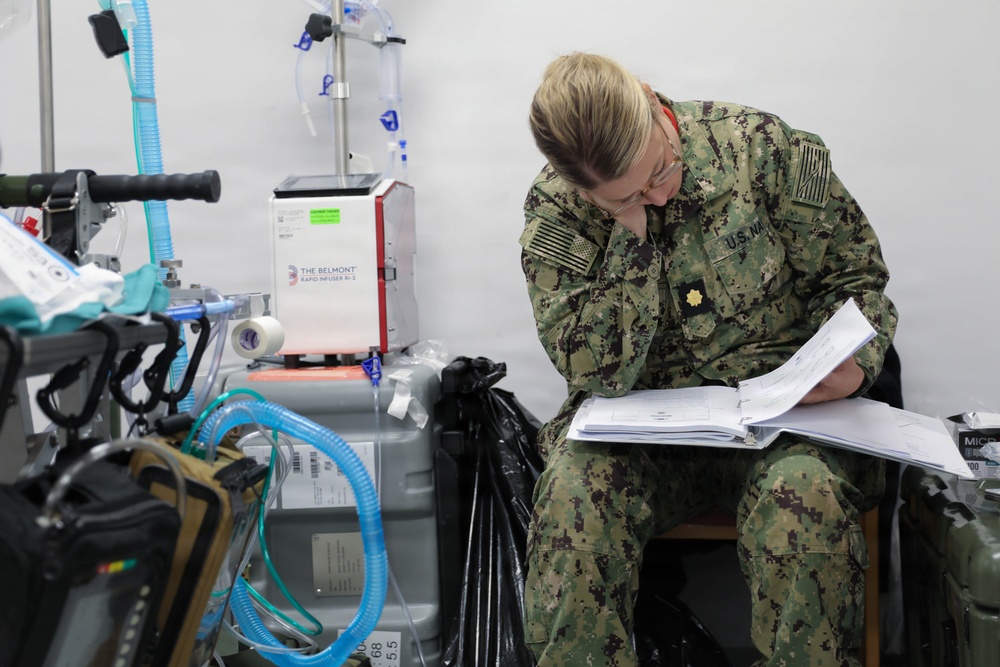A U.S. Navy service member in digital camouflage (NWU Type III) uniform is seated and reviewing documents in a binder within what appears to be a military medical facility or field hospital. The individual wears the rank insignia of a Lieutenant Commander (gold oak leaf) and glasses, and is intently focused on paperwork. Medical equipment including an IV stand and a Belmont Rapid Infuser RI-2 is visible in the background.