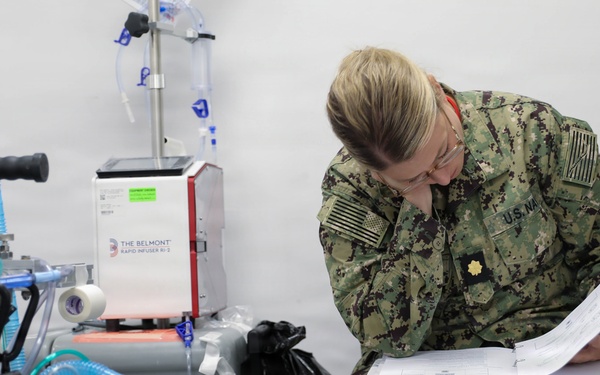 A U.S. Navy service member in digital camouflage (NWU Type III) uniform is seated and reviewing documents in a binder within what appears to be a military medical facility or field hospital. The individual wears the rank insignia of a Lieutenant Commander (gold oak leaf) and glasses, and is intently focused on paperwork. Medical equipment including an IV stand and a Belmont Rapid Infuser RI-2 is visible in the background.