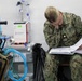 A U.S. Navy service member in digital camouflage (NWU Type III) uniform is seated and reviewing documents in a binder within what appears to be a military medical facility or field hospital. The individual wears the rank insignia of a Lieutenant Commander (gold oak leaf) and glasses, and is intently focused on paperwork. Medical equipment including an IV stand and a Belmont Rapid Infuser RI-2 is visible in the background.