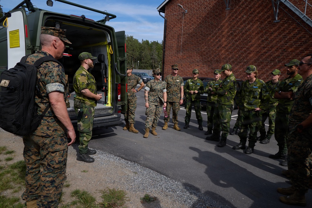 A group of military personnel in two distinct camouflage uniforms are gathered in a parking area next to a military van with its rear doors open. The group appears to include both US Marines (in MARPAT digital camouflage) and what appear to be Swedish Armed Forces soldiers (in Swedish M90 camouflage pattern). The setting appears to be a military installation, likely during a joint training exercise or exchange program.