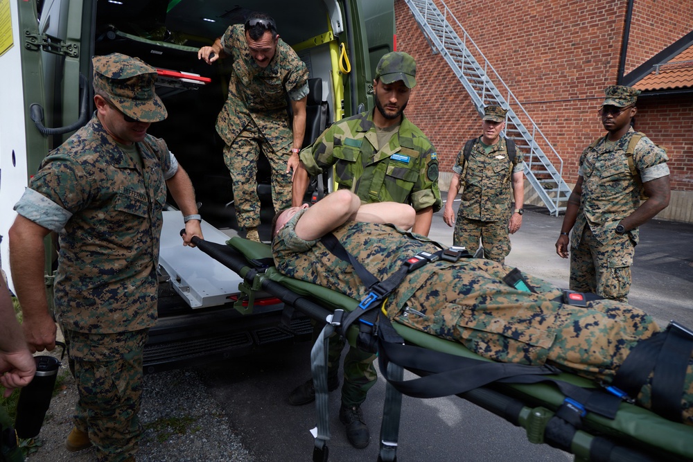 Military personnel in camouflage uniforms are loading or unloading a casualty on a stretcher into or out of an ambulance vehicle. The scene appears to be a medical training exercise or casualty evacuation drill, involving what appear to be U.S. Marines and at least one Swedish military soldier. The setting is an outdoor area adjacent to a brick building with an external metal staircase.