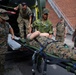 Military personnel in camouflage uniforms are loading or unloading a casualty on a stretcher into or out of an ambulance vehicle. The scene appears to be a medical training exercise or casualty evacuation drill, involving what appear to be U.S. Marines and at least one Swedish military soldier. The setting is an outdoor area adjacent to a brick building with an external metal staircase.