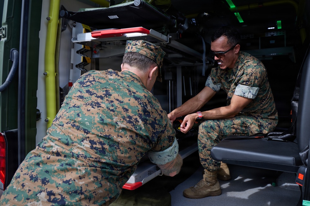 Two military personnel in MARPAT (Marine pattern) camouflage uniforms are inspecting or working on the interior of what appears to be a military ambulance or medical vehicle. One individual is crouching at the rear entrance while the other is seated inside the vehicle's cargo/patient compartment. They appear to be examining equipment or a mechanism near the vehicle's loading step.