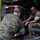 Two military personnel in MARPAT (Marine pattern) camouflage uniforms are inspecting or working on the interior of what appears to be a military ambulance or medical vehicle. One individual is crouching at the rear entrance while the other is seated inside the vehicle's cargo/patient compartment. They appear to be examining equipment or a mechanism near the vehicle's loading step.