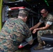 Two military personnel in MARPAT (Marine pattern) camouflage uniforms are inspecting or working on the interior of what appears to be a military ambulance or medical vehicle. One individual is crouching at the rear entrance while the other is seated inside the vehicle's cargo/patient compartment. They appear to be examining equipment or a mechanism near the vehicle's loading step.