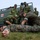 A military medical training exercise is taking place outdoors on a grass area. Personnel in two distinct camouflage uniforms — Swedish military pattern and US Marine Corps MARPAT — are practicing combat casualty care on a simulated casualty lying on the ground. A green military ambulance van marked with a Red Cross is visible in the background.