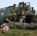 A military medical training exercise is taking place outdoors on a grass area. Personnel in two distinct camouflage uniforms — Swedish military pattern and US Marine Corps MARPAT — are practicing combat casualty care on a simulated casualty lying on the ground. A green military ambulance van marked with a Red Cross is visible in the background.
