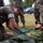 A military medical training exercise is taking place outdoors on a grass field. A service member in MARPAT digital camouflage uniform is providing medical treatment to a soldier lying on the ground wearing a different camouflage pattern (appears to be Swedish M90 woodland). Several other uniformed personnel are observing the procedure in the background.
