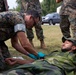 A military medical training exercise is taking place outdoors on a grass field. A service member in MARPAT digital camouflage uniform is providing medical treatment to a soldier lying on the ground wearing a different camouflage pattern (appears to be Swedish M90 woodland). Several other uniformed personnel are observing the procedure in the background.