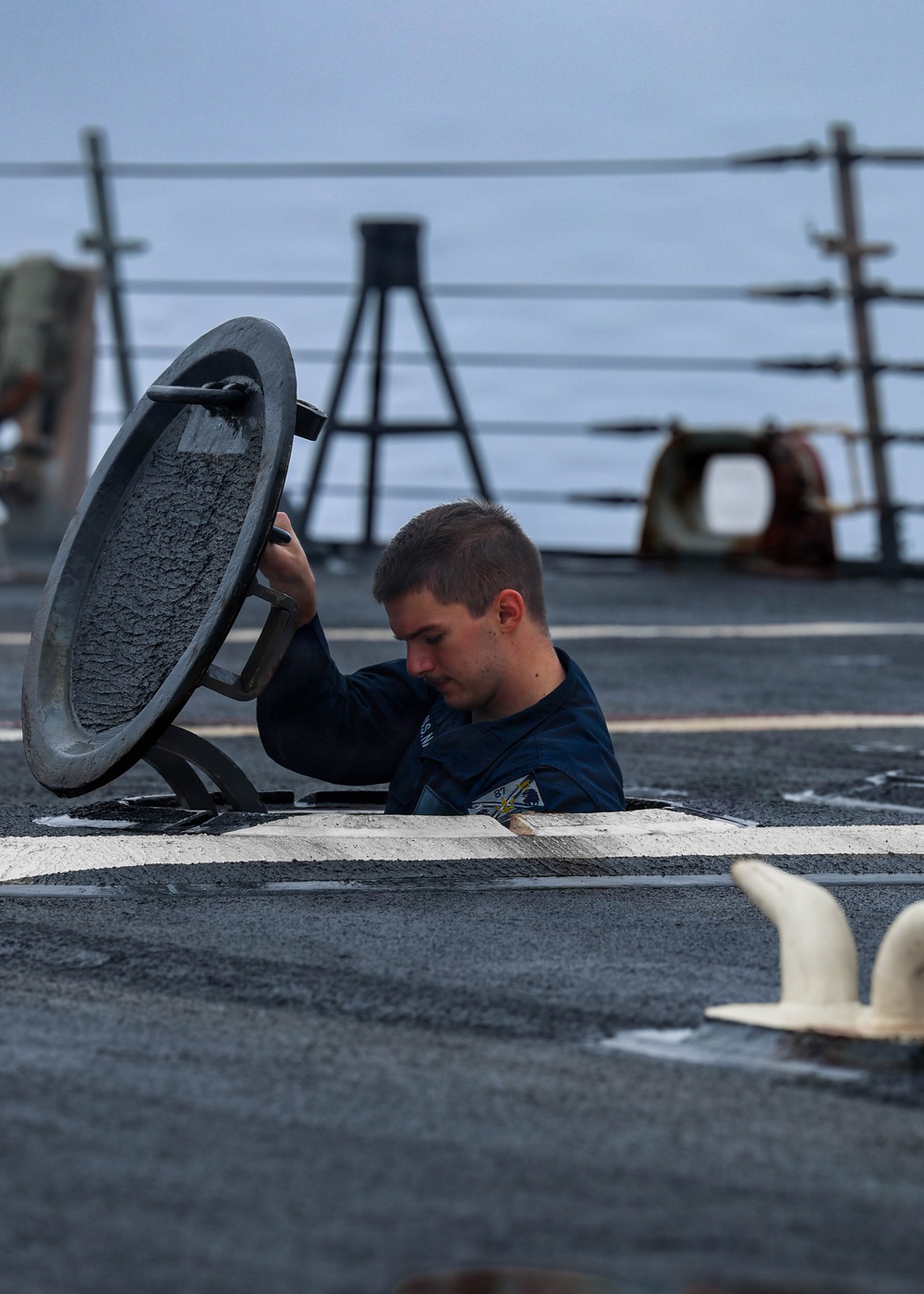 A young male sailor in a dark navy uniform is emerging from or descending into a deck hatch on what appears to be a naval vessel. He is lifting or holding open a heavy circular hatch cover while partially submerged in the opening. The image was taken on the ship's deck with the ocean and ship rigging visible in the background.