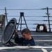 A young male sailor in a dark navy uniform is emerging from or descending into a deck hatch on what appears to be a naval vessel. He is lifting or holding open a heavy circular hatch cover while partially submerged in the opening. The image was taken on the ship's deck with the ocean and ship rigging visible in the background.
