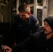 Two U.S. Navy personnel are working at a control panel aboard what appears to be a naval vessel. One sailor is wearing a standard U.S. Navy uniform with visible rank insignia, while the other is wearing a dark beanie and headset/hearing protection. They appear to be operating or monitoring equipment in a dimly lit shipboard compartment.
