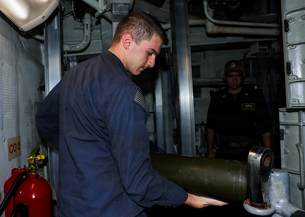 Two military personnel are working inside what appears to be a naval vessel's interior compartment. The foreground sailor in a blue coverall uniform is handling a large cylindrical olive-green object, likely an ordnance or equipment canister. A second sailor wearing a dark uniform and cap observes from a doorway in the background.