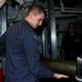 Two military personnel are working inside what appears to be a naval vessel's interior compartment. The foreground sailor in a blue coverall uniform is handling a large cylindrical olive-green object, likely an ordnance or equipment canister. A second sailor wearing a dark uniform and cap observes from a doorway in the background.