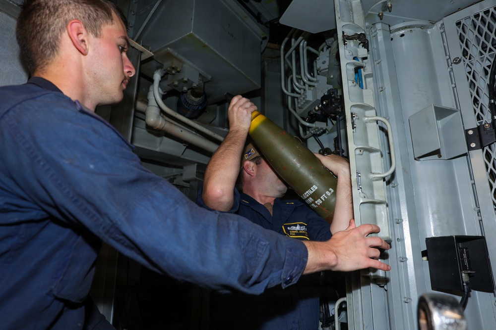 Two naval personnel in dark blue uniforms are handling a green cylindrical ordnance or projectile inside what appears to be a confined shipboard compartment. One sailor is wearing a name tag patch visible on their uniform. They appear to be loading or positioning the munition into a launcher or storage tube mounted on the bulkhead.