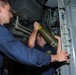 Two naval personnel in dark blue uniforms are handling a green cylindrical ordnance or projectile inside what appears to be a confined shipboard compartment. One sailor is wearing a name tag patch visible on their uniform. They appear to be loading or positioning the munition into a launcher or storage tube mounted on the bulkhead.