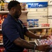 A man in a navy blue polo shirt with an emblem is stocking or organizing snack products on a display rack in what appears to be a military ship's store. The background signage reads 'YOUR SHIP YOUR STORE,' indicating this is likely a Navy ship's commissary or convenience store. The shelves behind him are stocked with various chip brands including Lay's, Fritos, Smartfood, and Doritos.