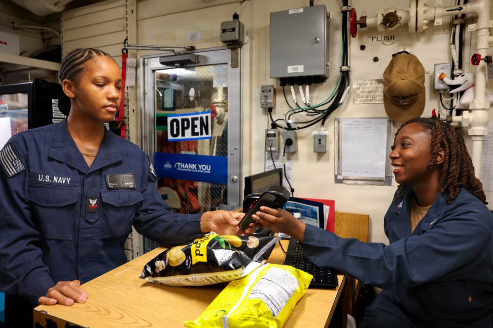 Two U.S. Navy servicewomen are conducting a transaction at a ship's store aboard a naval vessel. One sailor in uniform is making a purchase using a card reader while the other operates as the cashier. The setting is a compact onboard retail space with an 'OPEN' sign visible in the background.