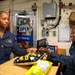 Two U.S. Navy servicewomen are conducting a transaction at a ship's store aboard a naval vessel. One sailor in uniform is making a purchase using a card reader while the other operates as the cashier. The setting is a compact onboard retail space with an 'OPEN' sign visible in the background.