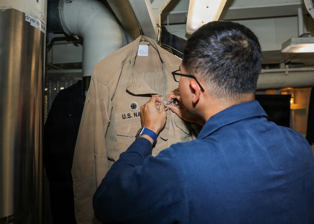 A person wearing glasses and a blue uniform is working on a tan/khaki U.S. Navy uniform shirt that is hanging on a rack. The individual appears to be attaching, adjusting, or removing insignia or fasteners from the shirt. The setting appears to be an interior space aboard a naval vessel, indicated by the industrial piping and equipment visible in the background.
