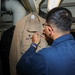 A person wearing glasses and a blue uniform is working on a tan/khaki U.S. Navy uniform shirt that is hanging on a rack. The individual appears to be attaching, adjusting, or removing insignia or fasteners from the shirt. The setting appears to be an interior space aboard a naval vessel, indicated by the industrial piping and equipment visible in the background.