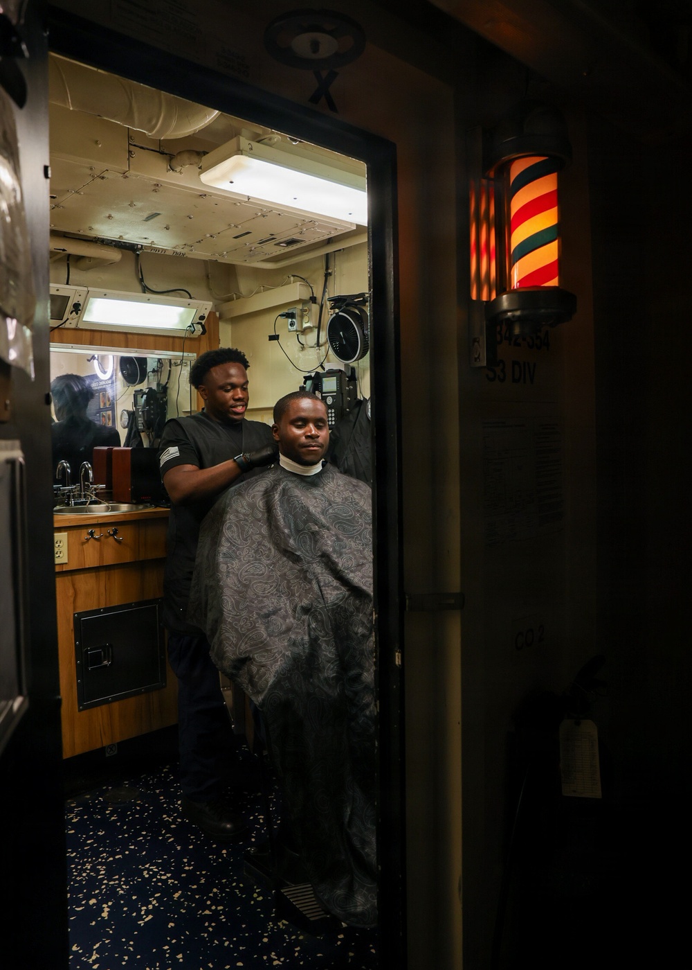A barber is cutting the hair of a seated client inside what appears to be a shipboard barbershop, likely aboard a U.S. Navy vessel. The client is draped in a patterned barber cape while the barber stands behind him working. A traditional illuminated barber pole is visible mounted on the wall outside the doorway.