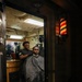 A barber is cutting the hair of a seated client inside what appears to be a shipboard barbershop, likely aboard a U.S. Navy vessel. The client is draped in a patterned barber cape while the barber stands behind him working. A traditional illuminated barber pole is visible mounted on the wall outside the doorway.