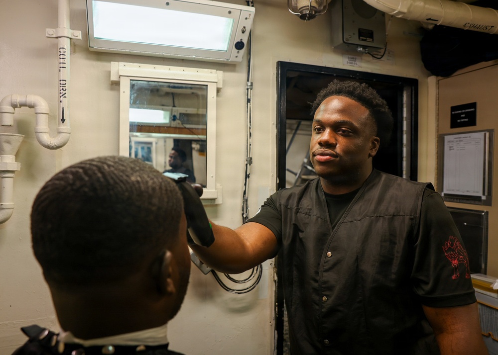 A barber is cutting the hair of a seated client in what appears to be a shipboard barbershop, likely aboard a naval vessel. The barber is wearing a dark vest with a red graphic design on the sleeve and is holding electric clippers. The utilitarian environment, with visible pipes, metal doors, and posted notices, is consistent with a military ship interior.