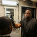 A barber is cutting the hair of a seated client in what appears to be a shipboard barbershop, likely aboard a naval vessel. The barber is wearing a dark vest with a red graphic design on the sleeve and is holding electric clippers. The utilitarian environment, with visible pipes, metal doors, and posted notices, is consistent with a military ship interior.