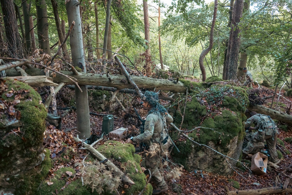 The image shows military personnel in full combat gear operating in a dense woodland environment. At least three soldiers are visible navigating through rocky, moss-covered terrain with fallen trees and leaf-covered ground. They appear to be conducting a field training exercise or tactical operation, carrying equipment and supplies.